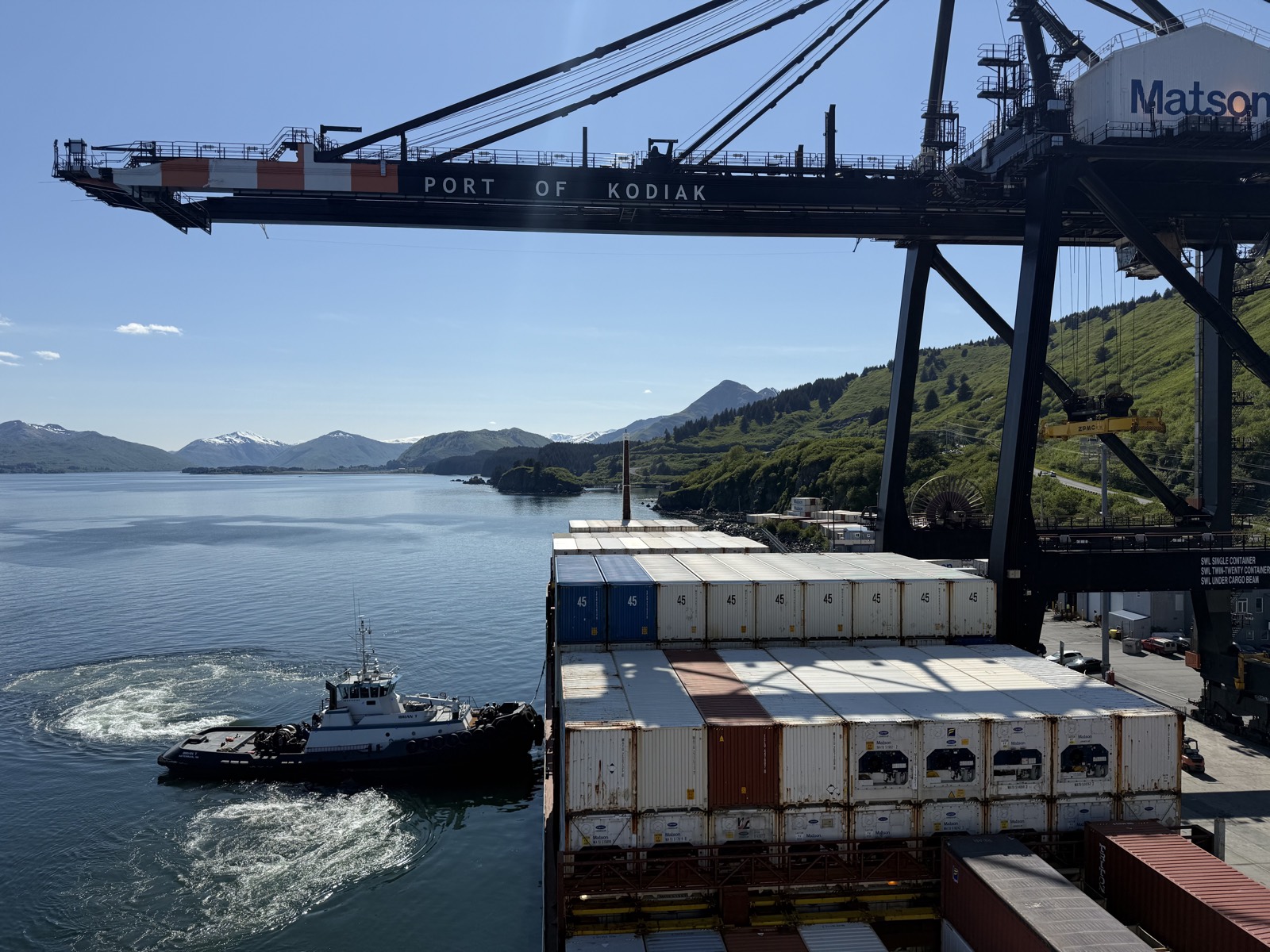 A container ship at the Port of Kodiak with a tug and green island hills