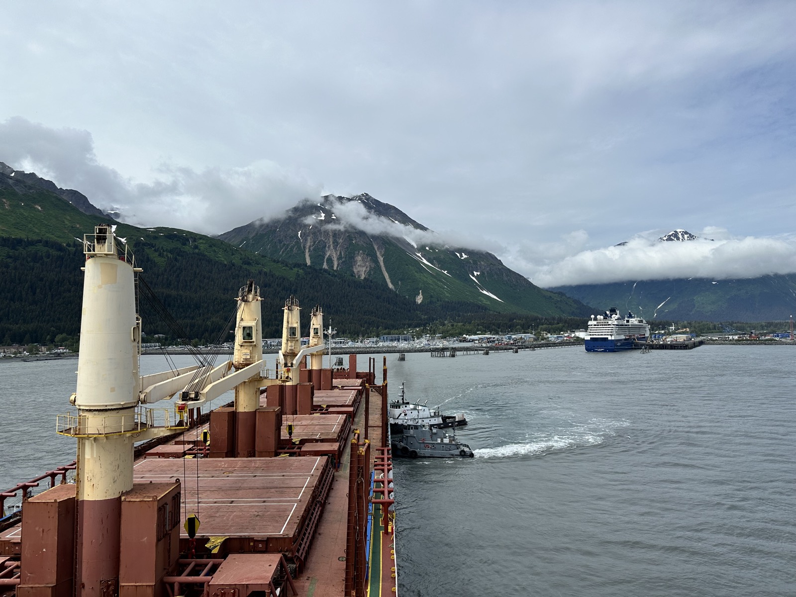 A cargo ship approaching the port of Seward in Resurrection Bay, with a cruise ship and green mountains